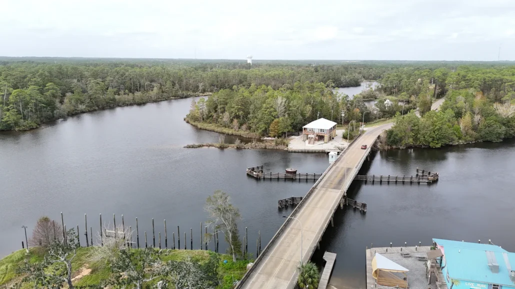 Drone view along a low two-lane bridge over a winding coastal river, with fender dolphins at the channel span