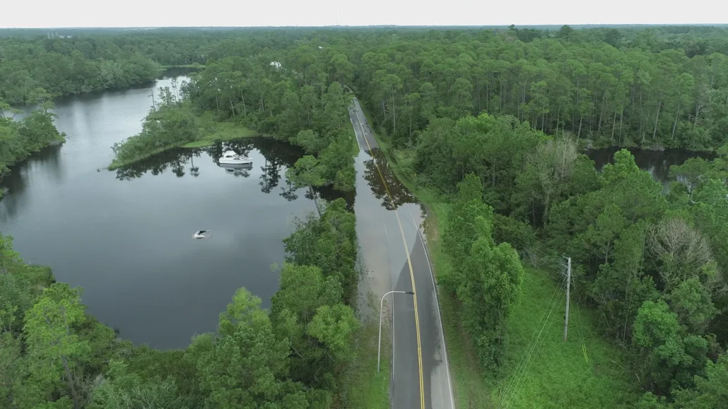 Floodwater covering a two-lane road through a forested river floodplain, with a boat on the water