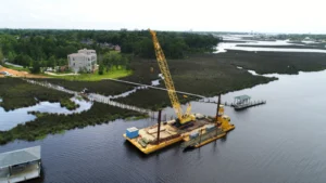Aerial view of a barge-mounted crane driving timber piles for a residential pier across a coastal salt marsh