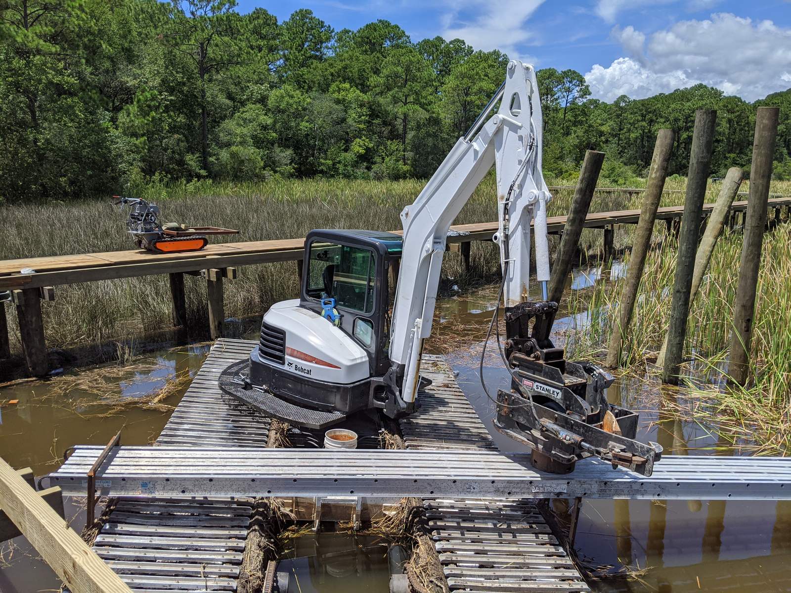 Timber pier under construction across a tidal marsh, with a marsh buggy excavator working