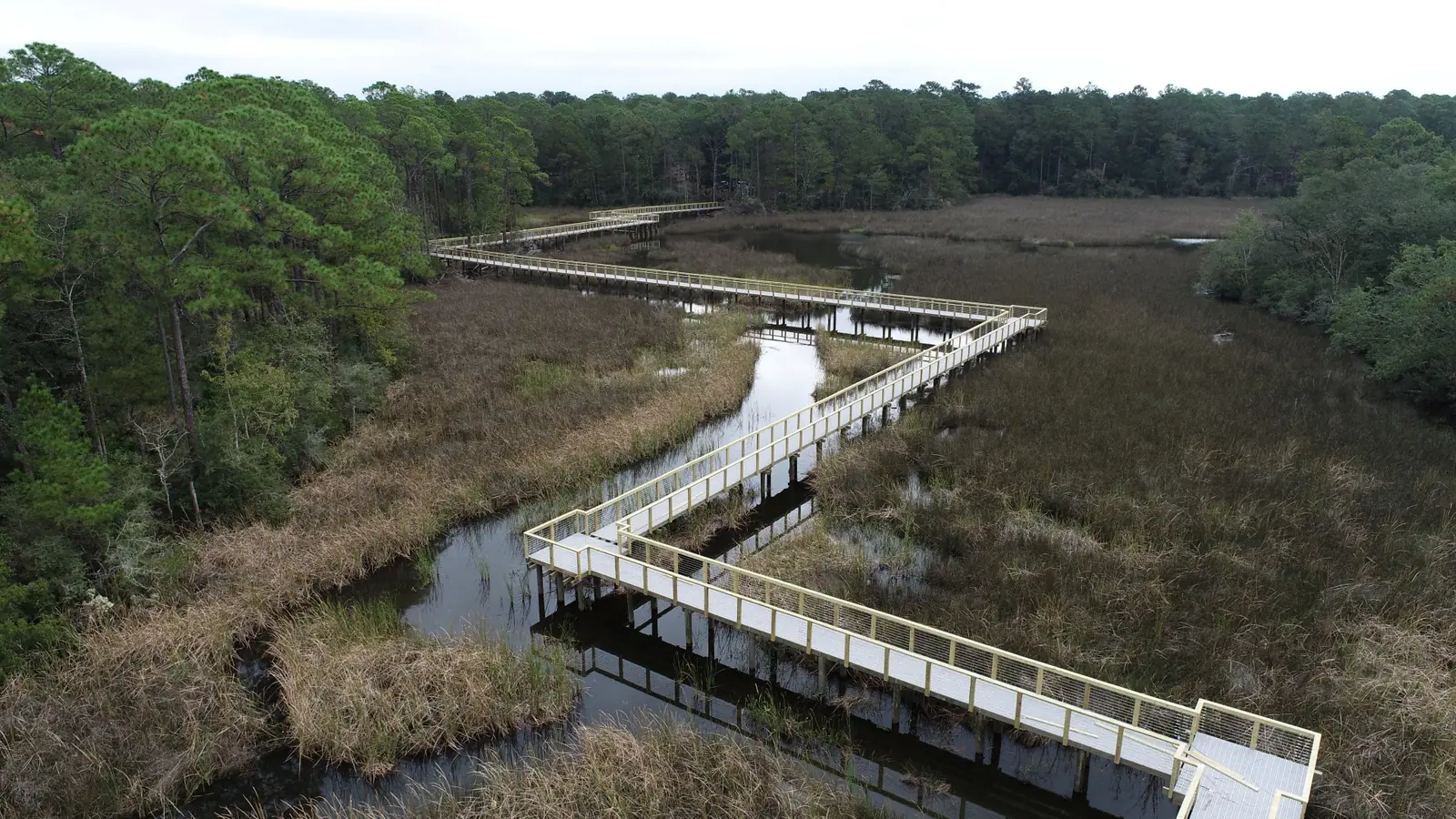 Low aerial view along a zigzag marsh boardwalk, with the deck and railings reflected in the water below