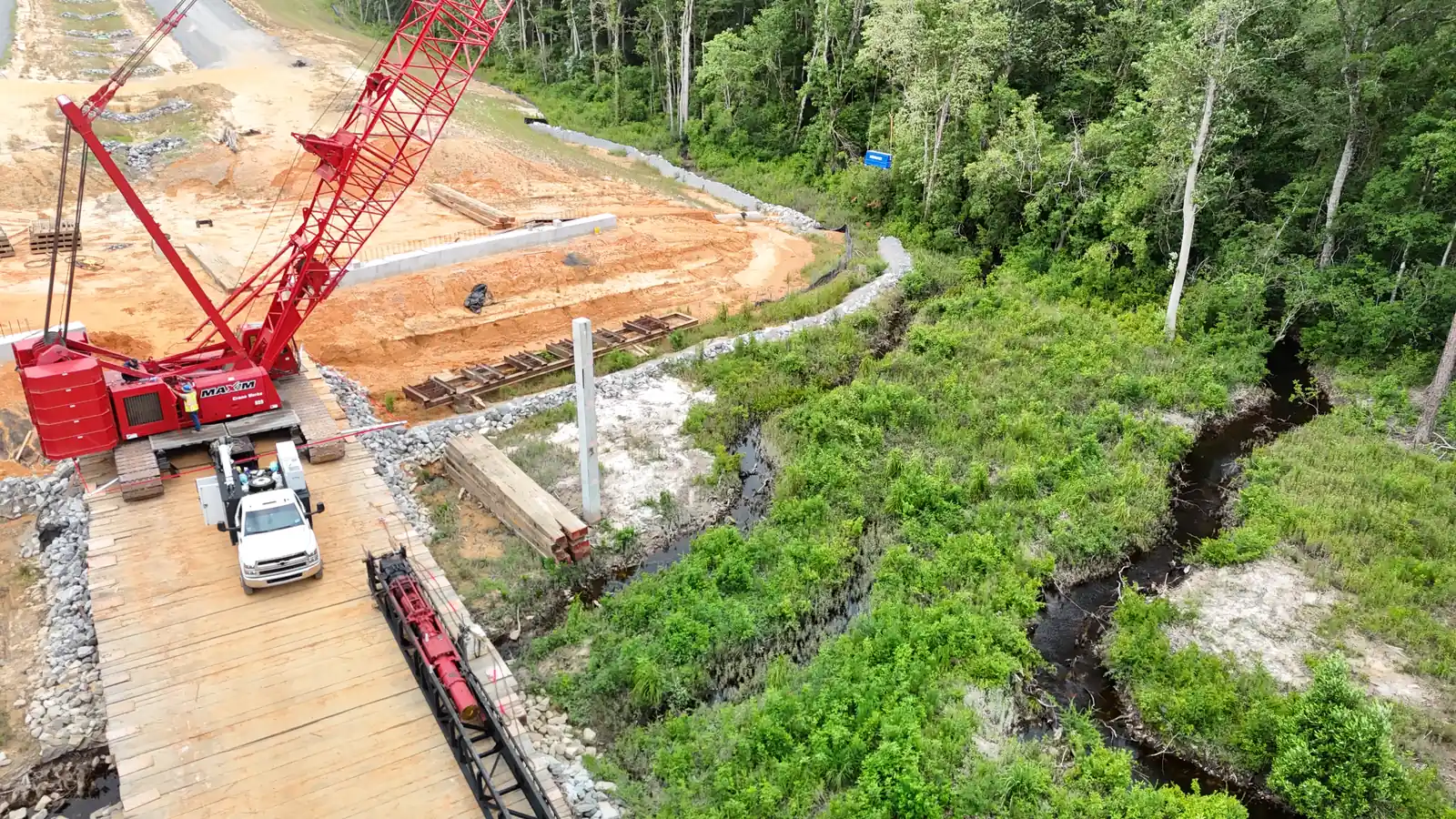 Drone view of a red crawler crane and a pickup truck on a timber work trestle over a blackwater stream and forested wetland during SR 57 construction in Mississippi, with riprap and a staging area