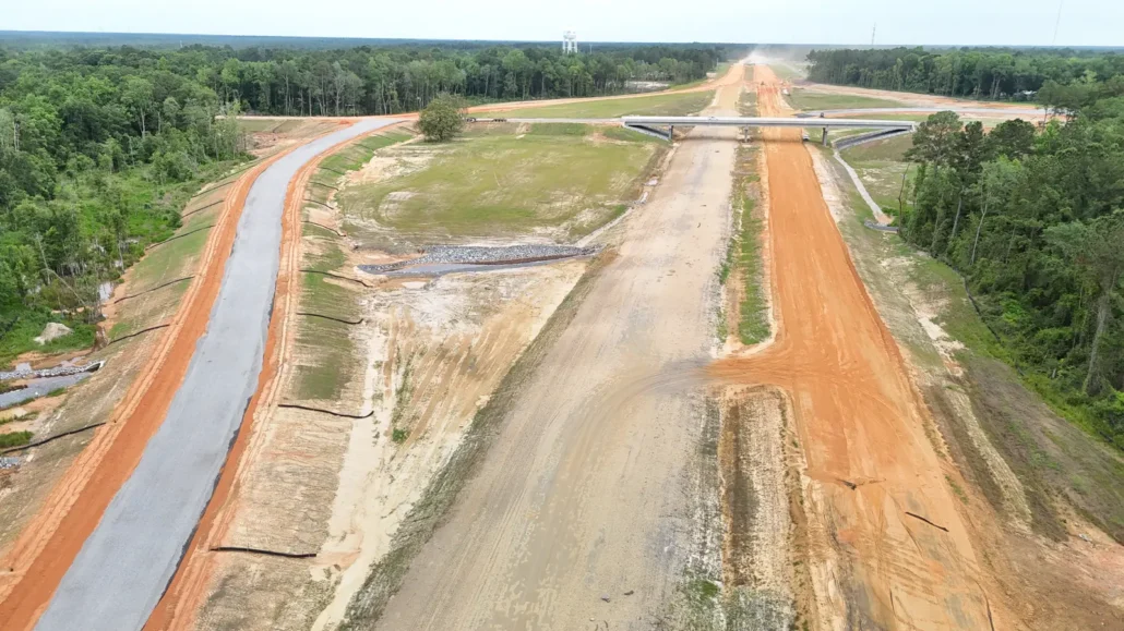 New highway corridor under construction, with graded lanes, silt fence and riprap along the edges and an overpass in the distance