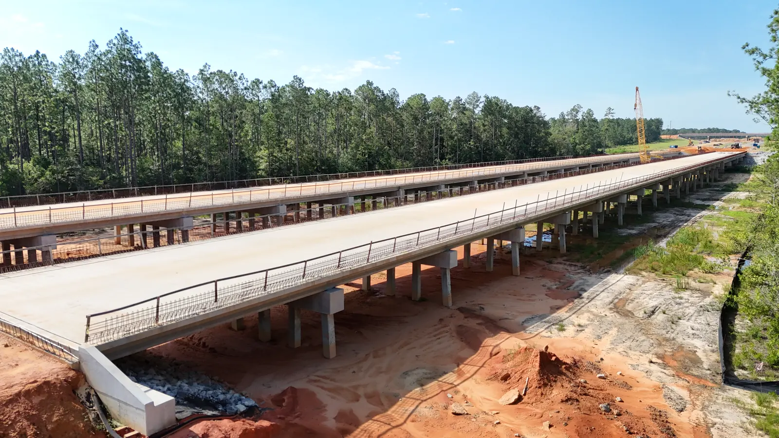 Drone view of new twin concrete bridge decks on pile bents under construction on SR 57 in Mississippi, with cleared sandy ground below and pine forest on both sides