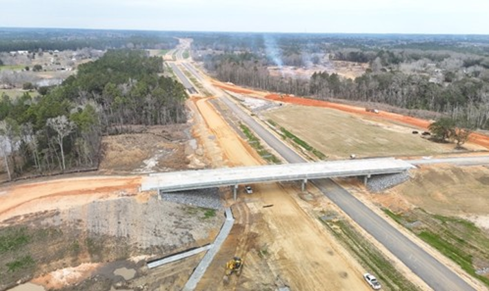 Aerial view of a highway overpass under construction, with graded and stabilized embankment slopes and riprap-lined drainage channels.
