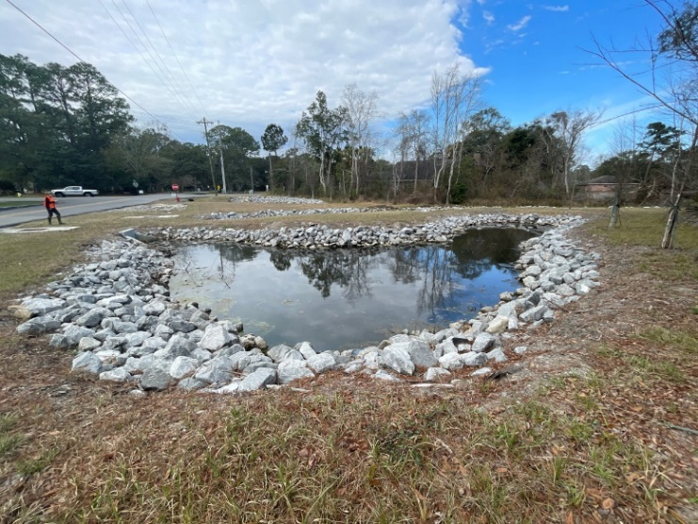 Stormwater pond lined with riprap beside a two-lane roadway, with grassed banks and trees behind it.