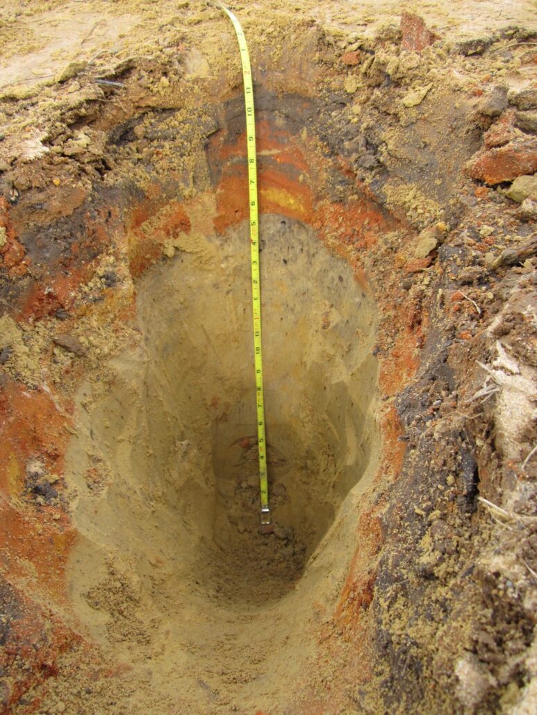 Deep soil pit in sandy soil with dark and orange-red bands over pale sand, tape for scale
